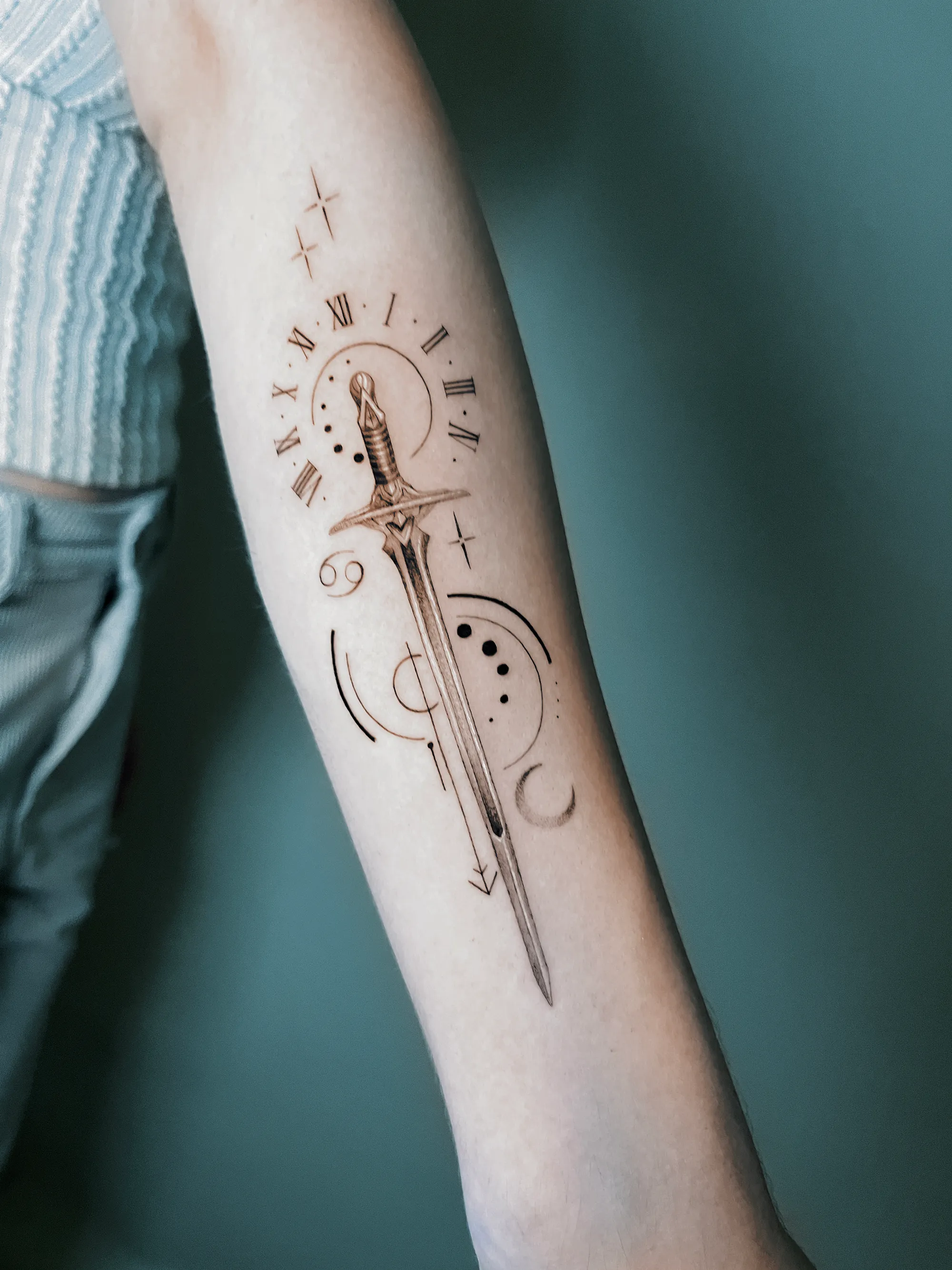 Fine line sword forearm tattoo with celestial elements, Roman numerals, geometric linework, and delicate black and grey shading.